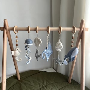 以下が含まれることがあります： 木製のベビージム。海をテーマにしたおもちゃが吊り下げられています。おもちゃには、タツノオトシゴ、クラゲ、貝殻、マンタ、ヒトデ、クジラが含まれます。おもちゃは白、青、グレーの色合いです。ジムは緑色のキルトの表面に置かれています。