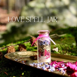 May include: A small glass jar with a pink ribbon tied around the top, filled with dried herbs, flowers, and crystals. The jar is sitting on a gold tray with rose petals and lavender flowers scattered around it. The text "LOVE SPELL JAR" is written in white letters on a blurred background.