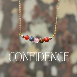 May include: A gold chain necklace with a row of six colorful gemstone beads. The beads are red, green, pink, brown, and red. The necklace has the word "CONFIDENCE" written below it.
