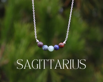 May include: A silver chain necklace with five gemstone beads in shades of red, blue, and green. The beads spell out "SAGITTARIUS" in the image.