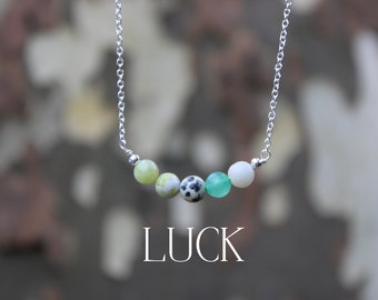 May include: A silver chain necklace with five green and white gemstone beads. The beads are arranged in a curved line and the word "LUCK" is printed below the necklace.
