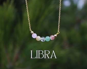 May include: A gold chain necklace with a row of pastel-colored beads, including pink, yellow, clear, green, and pink. The text "LIBRA" is written below the necklace.
