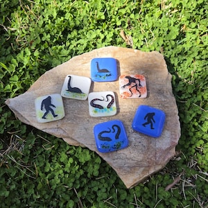 May include: Seven square glass tiles with various monster illustrations, including the Loch Ness Monster and Bigfoot, are displayed on a stone. The tiles feature blue, white, and orange backgrounds with black and green designs. The tiles are set against a backdrop of green clover.