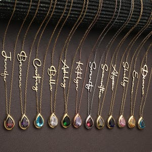 May include: A collection of gold and silver name necklaces with teardrop-shaped gemstone pendants. Each necklace features a different name in a cursive font. The gemstones are various colors, including red, purple, and blue.
