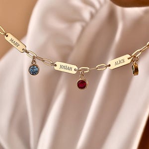 May include: Gold-toned necklace with rectangular nameplates and gemstone charms. The nameplates read "MARY", "JOSIAH", and "ALICE". The charms are blue, red, and green. The necklace is displayed against a soft, white background.