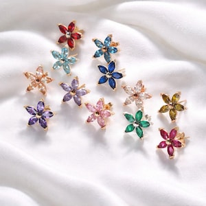 May include: A collection of flower-shaped stud earrings in various colors, including red, blue, purple, pink, and green. Each earring features a gold-colored base and is set with multiple gemstones. The earrings are displayed on a white, textured surface.