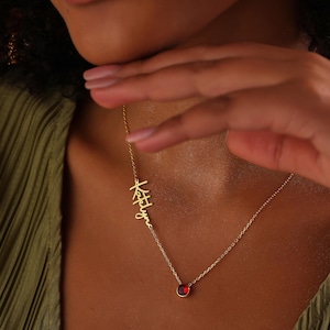 May include: A gold-colored necklace with a pendant featuring a name in cursive script and a small, round, red gemstone. The necklace is worn against a green top.