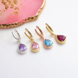 May include: Four pairs of teardrop earrings in silver, gold, and rose gold tones. Each earring features a different colored gemstone: purple, pink, blue, and red. The earrings are displayed on a white surface.