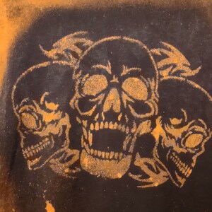 Puede incluir: Una sudadera negra con un diseño salpicado de lejía. El diseño presenta tres calaveras negras con ojos y dientes blancos. Las calaveras están dispuestas en forma triangular.