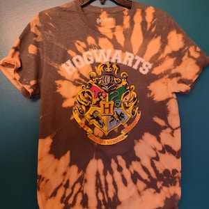 May include: A gray tie-dye t-shirt with the Hogwarts crest and the text "HOGWARTS" in white. The crest features a lion, eagle, badger, and snake.