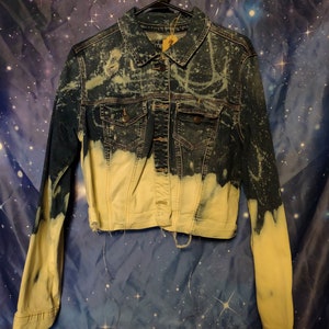 May include: A blue denim jacket with a bleach-splattered design. The jacket has a yellowed appearance, especially on the sleeves and bottom hem. The jacket has a button closure and two front pockets.