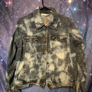 May include: A blue denim jacket with a white bleach splatter pattern. The jacket has a button closure and two front pockets.