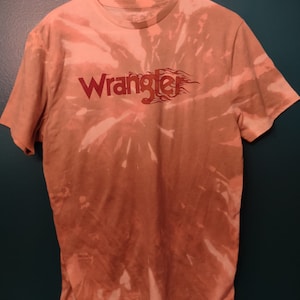 May include: A tie-dye t-shirt in shades of orange and white with the word "Wrangler" in red lettering with a flame design.