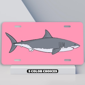 May include: A pink license plate with a white great white shark illustration. The shark is facing right and has a gray body with a white underbelly. The text "5 COLOR CHOICES" is printed in black at the bottom of the plate.