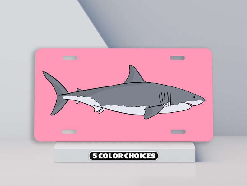 May include: A pink license plate with a white great white shark illustration. The shark is facing right and has a gray body with a white underbelly. The text "5 COLOR CHOICES" is printed in black at the bottom of the plate.