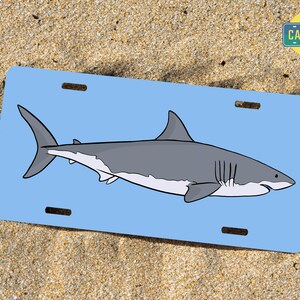 May include: A blue and white license plate featuring a cartoon illustration of a great white shark. The text "The Car Bar" is in a white rectangle in the top right corner of the plate.