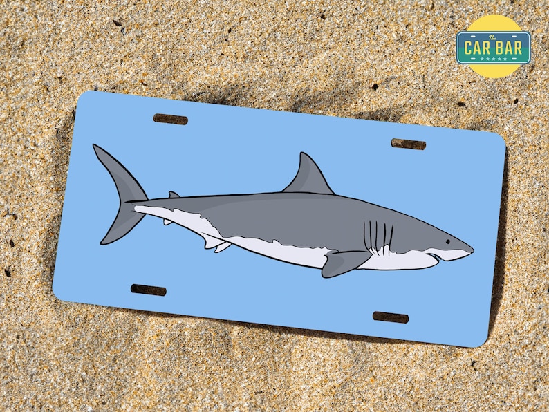 May include: A blue and white license plate featuring a cartoon illustration of a great white shark. The text "The Car Bar" is in a white rectangle in the top right corner of the plate.