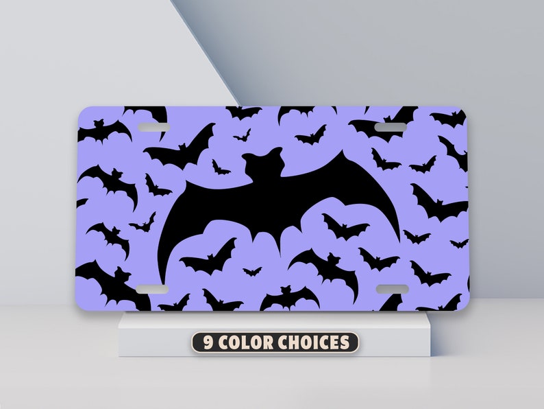May include: A purple license plate with a black bat silhouette pattern. The bat silhouette is repeated throughout the plate. The plate has a hole in the top center and two holes on the bottom corners.