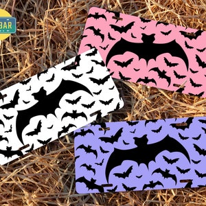 May include: Three different colored license plates with a black bat pattern. The plates are white, pink, and purple. The bats are all the same size and shape, and they are arranged in a random pattern. The text "The Car Bar" is on the white plate.