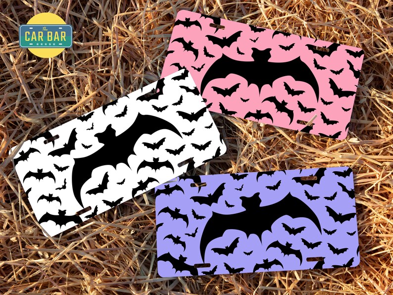 May include: Three different colored license plates with a black bat pattern. The plates are white, pink, and purple. The bats are all the same size and shape, and they are arranged in a random pattern. The text "The Car Bar" is on the white plate.