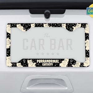 Puede incluir: Marco de matr&iacute;cula negro y blanco con un patr&oacute;n repetitivo de gatos blancos. El marco tiene el texto "The Car Bar" y "Purranormal Cattivity" impreso en &eacute;l.