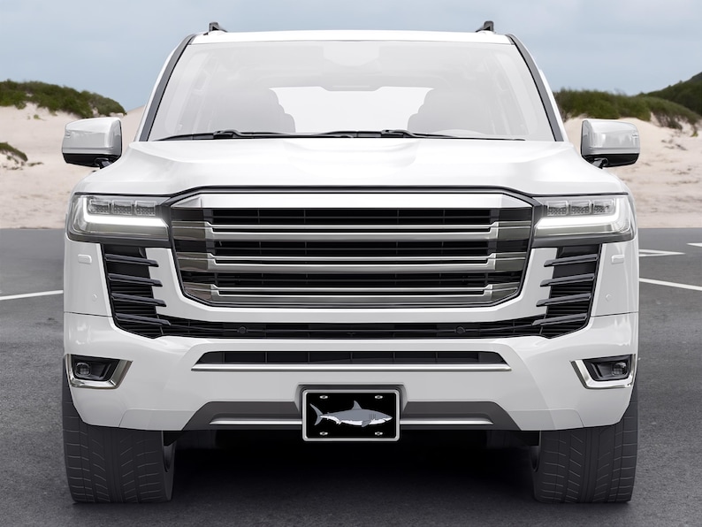 May include: White SUV with a black grille and a shark license plate. The vehicle is parked on a paved road with sand dunes in the background.