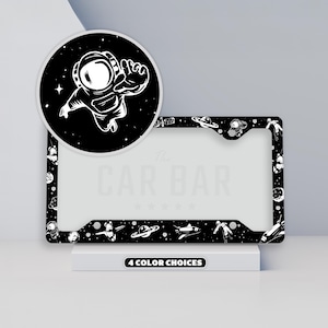 May include: Black license plate frame with a space theme. The frame features a repeating pattern of planets, astronauts, and spaceships. A circular graphic shows a white astronaut on a black background. The text "The CAR BAR" is visible. The bottom of the frame reads "4 COLOR CHOICES."
