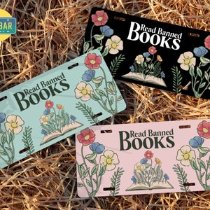 May include: Three different license plate designs with the text "Read Banned Books" and an open book with flowers growing out of it. The license plates are blue, black, and pink.