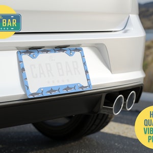 May include: A blue license plate frame with a shark pattern. The frame is attached to the back of a white car. The text "The Car Bar" is printed on the frame.