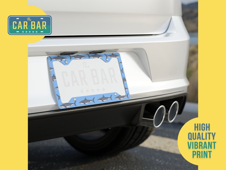 May include: A blue license plate frame with a shark pattern. The frame is attached to the back of a white car. The text "The Car Bar" is printed on the frame.