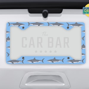 May include: A blue license plate frame with a white shark pattern and the text "The Car Bar" in white.