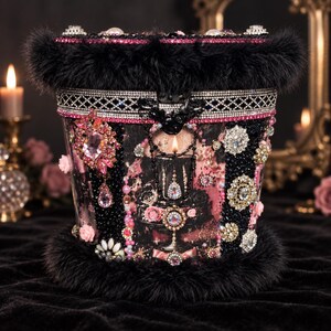 Könnte beinhalten: Ein zylindrischer Behälter, der oben und unten mit schwarzem Kunstfellbesatz verziert ist. Die Außenseite zeigt eine Collage aus rosa Blumenmotiven, schwarzen Perlenpaneelen, silbernen Strassbändern sowie verschiedenen Kristallbroschen und Perlen.