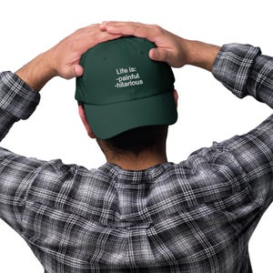 May include: A dark green baseball cap with white text that reads "Life is: -painful -hilarious".