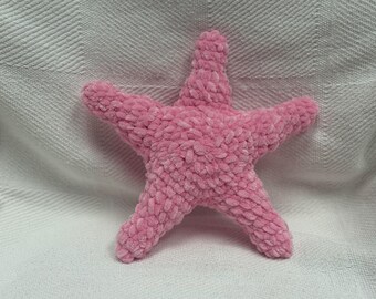 Puede incluir: Un juguete de estrella de mar rosa de felpa con cinco puntas. El material suave y texturizado es de un tono rosa claro. El juguete descansa sobre una superficie blanca y texturizada.