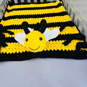 May include: A crocheted baby blanket with a bee design. The blanket is yellow and black striped with a 3D bee in the center. The bee has a yellow body, black eyes, and white wings.