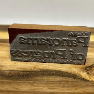 Pode incluir: Um carimbo de madeira com a parte superior vermelha e uma base de metal. A base de metal tem as palavras "Of Progress" e "Panorama" gravadas.