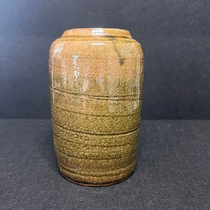 Puede incluir: Un jarrón cilíndrico de cerámica con una superficie texturizada y moteada. El jarrón tiene una combinación de colores marrón y verde oliva con líneas horizontales grabadas alrededor del cuerpo. El borde superior es de color marrón oscuro. El jarrón está sobre una superficie oscura.