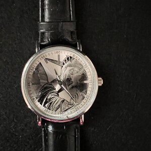 May include: A silver wristwatch with a black leather band. The watch face features a detailed engraving of the Statue of Liberty.