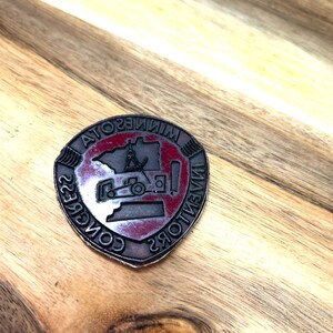 Pode incluir: Um broche de metal vermelho, branco e preto com as palavras "Inventors Congress" e "Minnesota" ao redor da borda. O broche apresenta uma imagem estilizada do estado de Minnesota com um trator dentro.