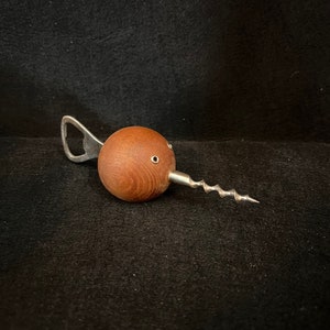 May include: A wooden bottle opener with a corkscrew and a bottle cap opener. The opener is shaped like a bird's head with two small holes for eyes.