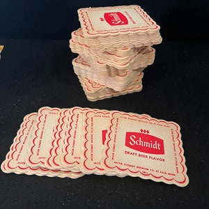 May include: A stack of brown cardboard coasters with a red logo that says "Schmidt Draft Beer Flavor" and "Jacob Schmidt Brewing Co., St. Paul, Minn."