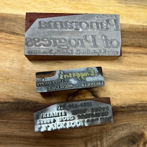 Pode incluir: Três blocos de impressão de metal vintage. O bloco superior é de madeira marrom com uma placa de metal que diz "of Progress". O bloco do meio diz "LAKEVILLE - PRIOR LAKE LADIES SHOPPE - Emporium's". O bloco inferior diz "PICK LOOP CO. NORTHLAND BOBBY DODD LAKEVILLE".