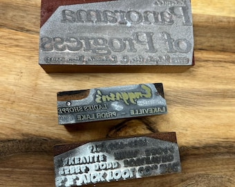 May include: Three vintage metal printing blocks. The top block is brown wood with a metal plate that reads "of Progress". The middle block reads "LAKEVILLE - PRIOR LAKE LADIES SHOPPE - Emporium's". The bottom block reads "PICK LOOP CO. NORTHLAND BOBBY DODD LAKEVILLE".