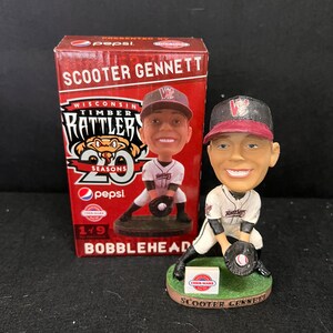 Könnte beinhalten: Eine rot-weiß-schwarze Bobblehead-Figur eines Baseballspielers, der ein Trikot der Wisconsin Timber Rattlers trägt. Die Figur hält einen Baseballhandschuh und steht auf einem grünen Sockel. Der Sockel trägt ein kleines rot-weißes "Cher-Make"-Logo und die Worte "Scooter Gennett". Die Figur ist in einer roten Box verpackt, auf der "Scooter Gennett" und "Wisconsin Timber Rattlers" steht.