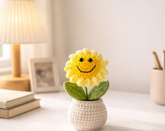 May include: A crocheted sunflower with a smiling face in a small, off-white crocheted pot. The sunflower has yellow petals, green leaves, and a black embroidered smile. The pot is sitting on a white desk.