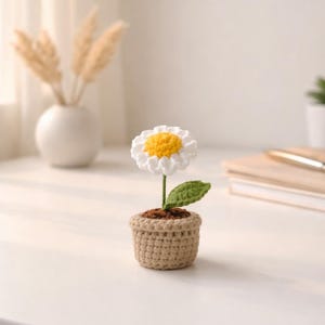 Op de afbeelding: Een gehaakte madeliefbloem met witte bloemblaadjes en een gele kern, geplant in een kleine, beige gehaakte pot. De bloem heeft een groene stengel en blad. De pot is gevuld met bruin materiaal, dat op aarde lijkt.