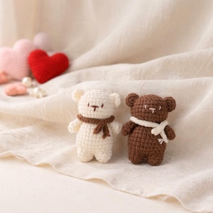 May include: Two small, handmade crochet teddy bears. One is white with a brown scarf, the other is brown with a white scarf. They have small black embroidered details. The bears are on a cream-colored fabric, with crochet hearts in the background.