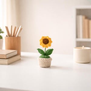 May include: A handcrafted crochet sunflower in a small pot. The flower has yellow petals and a brown center, with green leaves and a beige pot. The sunflower is placed on a white surface.