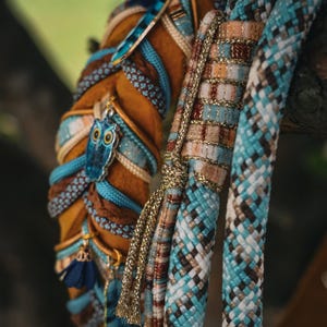 Könnte beinhalten: Nahaufnahme mehrerer Hundehalsbänder mit aufwendigen Designs. Die Halsbänder weisen eine Mischung aus braunen, blauen und goldenen Farben auf, mit geflochtenem Seil und dekorativen Anhängern, darunter ein Eulenanhänger und Federn. Die Halsbänder hängen in einer natürlichen Umgebung.