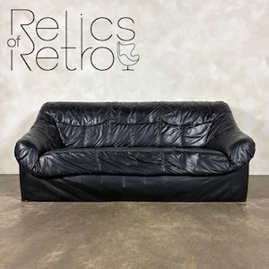 May include: Black leather sofa with a tufted design. The sofa has a low profile and a rectangular shape. The image includes the text "Relics of Retro" in a stylized font above the sofa.